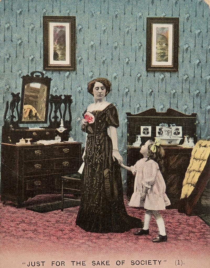 woman in Edwardian evening dress and gloves touches hand of small girl clinging to skirt as they stand in front of a dressing table and dresser
